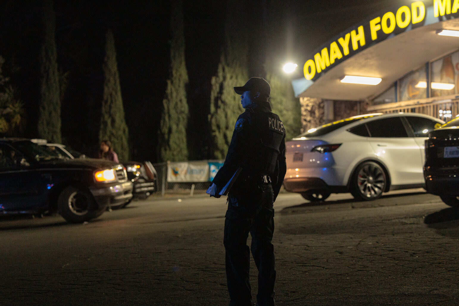 A nighttime scene in front of the “Omayh Food Mart & Liquor” storefront. A uniformed police officer stands in profile near a white vehicle, wearing a dark cap and vest labeled “POLICE.” Tall trees form a dark backdrop, and bright overhead lights illuminate the store’s entrance.