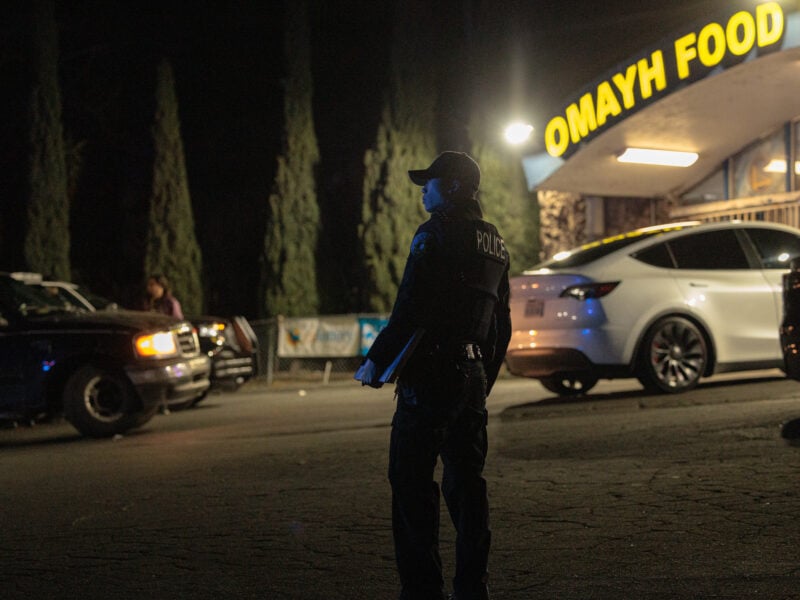 A nighttime scene in front of the “Omayh Food Mart & Liquor” storefront. A uniformed police officer stands in profile near a white vehicle, wearing a dark cap and vest labeled “POLICE.” Tall trees form a dark backdrop, and bright overhead lights illuminate the store’s entrance.