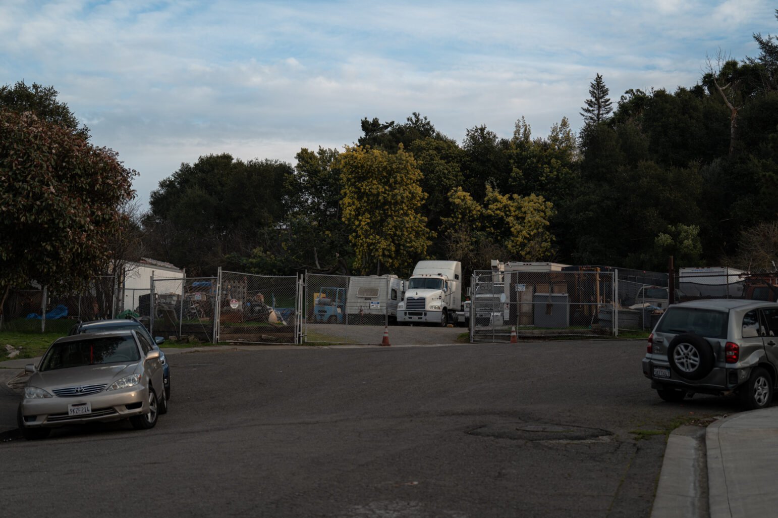 A fenced‐off lot at the end of a residential street, containing several large vehicles including a white semi‐truck and various equipment. Cars are parked along the curb on either side, and trees with green foliage rise behind the lot under a partly cloudy sky.