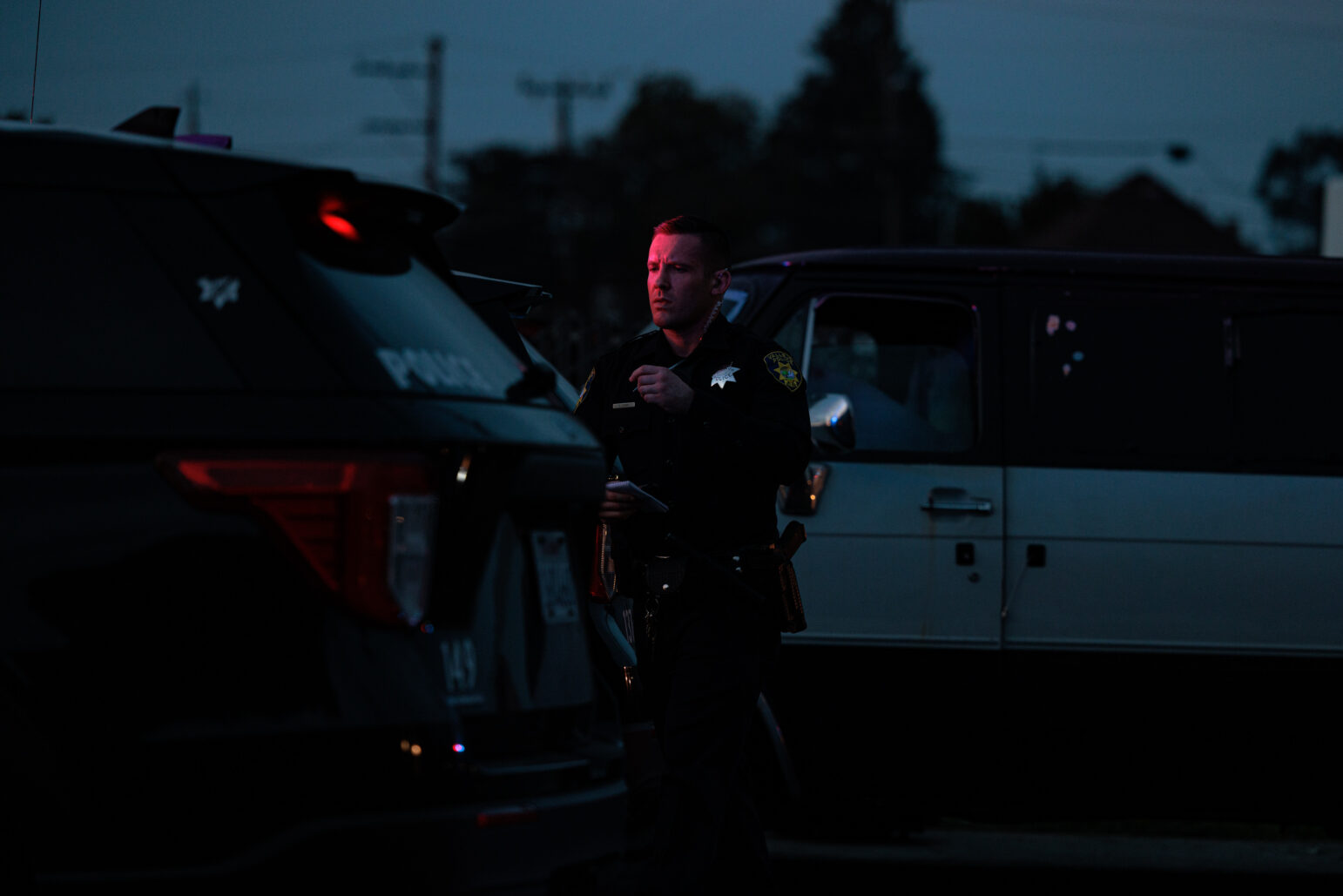 A dimly lit scene at dusk. A uniformed police officer stands beside a black patrol SUV, his face illuminated by red emergency lights. Behind him, a two-tone van is partially visible. Silhouetted trees and utility poles appear in the background.