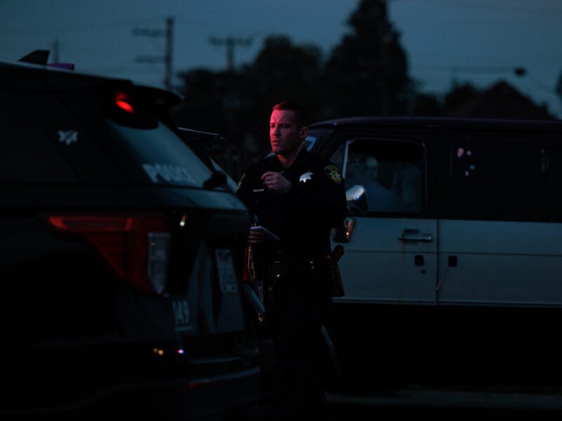 A dimly lit scene at dusk. A uniformed police officer stands beside a black patrol SUV, his face illuminated by red emergency lights. Behind him, a two-tone van is partially visible. Silhouetted trees and utility poles appear in the background.