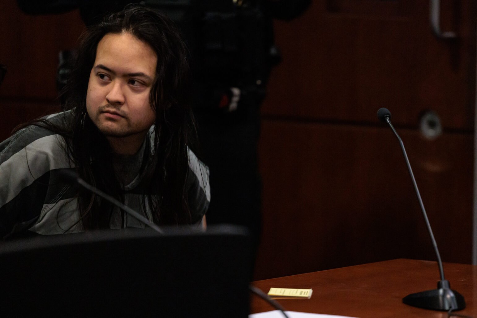 A male individual with long, dark hair sits in a courtroom wearing a gray-striped jail uniform. He looks to his left with a neutral or slightly alert expression. A wooden desk and microphones appear in the foreground, and the background is softly out of focus.