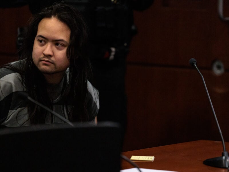 A male individual with long, dark hair sits in a courtroom wearing a gray-striped jail uniform. He looks to his left with a neutral or slightly alert expression. A wooden desk and microphones appear in the foreground, and the background is softly out of focus.