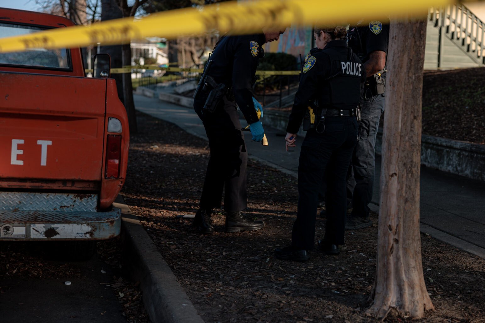 An orange pickup truck with chipped paint is parked at the curb. Yellow police tape crosses in front of the frame. Two police officers in dark uniforms and boots stand near the rear of the truck, leaning down as if examining something. One officer is wearing bright blue protective gloves and holding a recovered revolver.