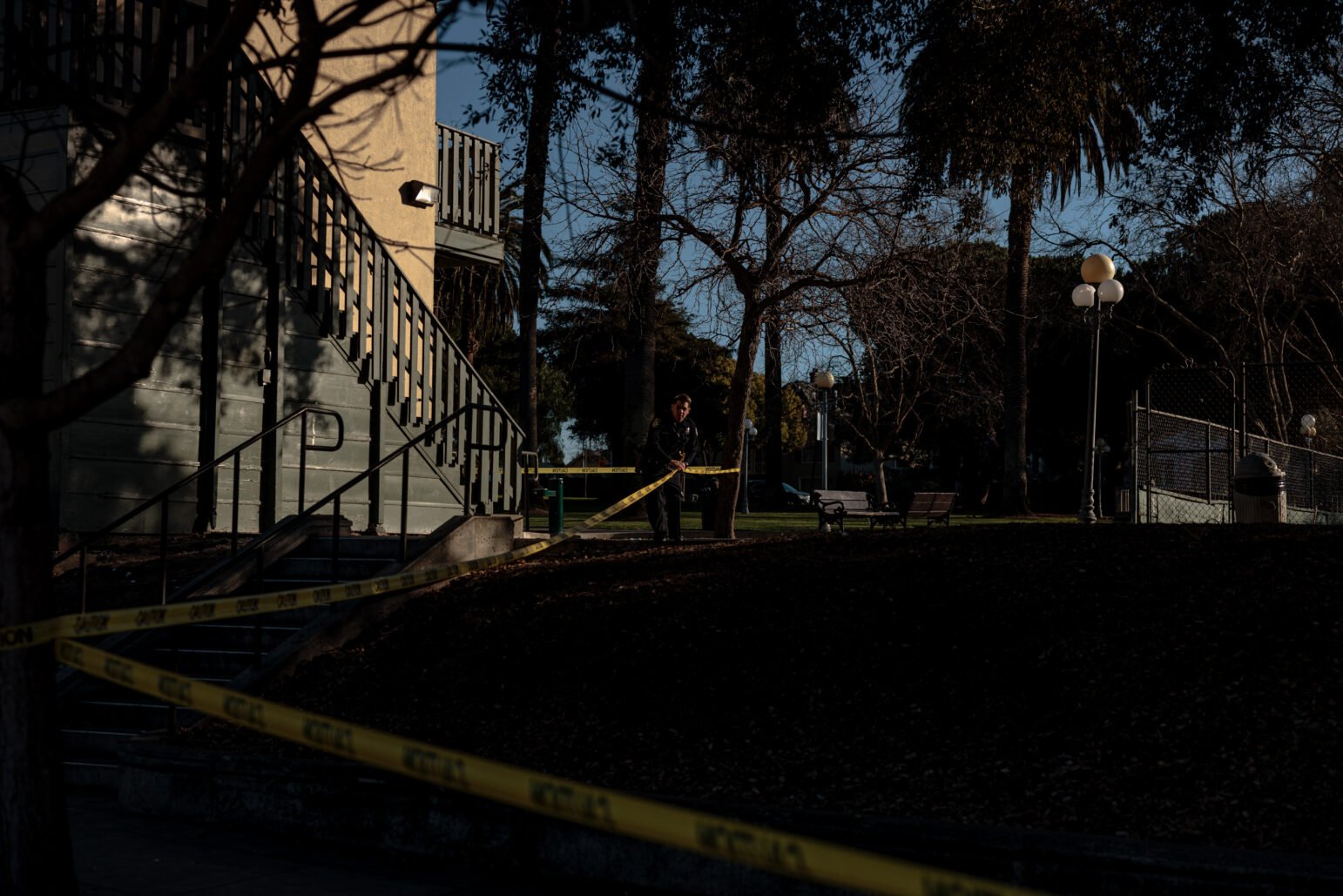 A wider shot of a park scene cordoned off by yellow police tape. A set of outdoor stairs attached to a light-colored building is visible on the left, with a police officer standing in the distance near the tape. The surrounding area has leaf-covered ground, trees, and lampposts, suggesting the scene is in a public park.