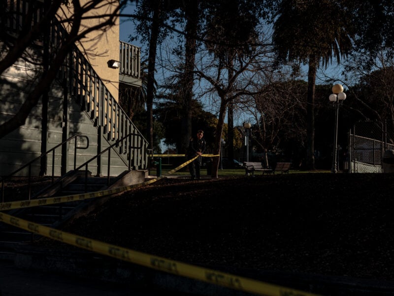 A wider shot of a park scene cordoned off by yellow police tape. A set of outdoor stairs attached to a light-colored building is visible on the left, with a police officer standing in the distance near the tape. The surrounding area has leaf-covered ground, trees, and lampposts, suggesting the scene is in a public park.