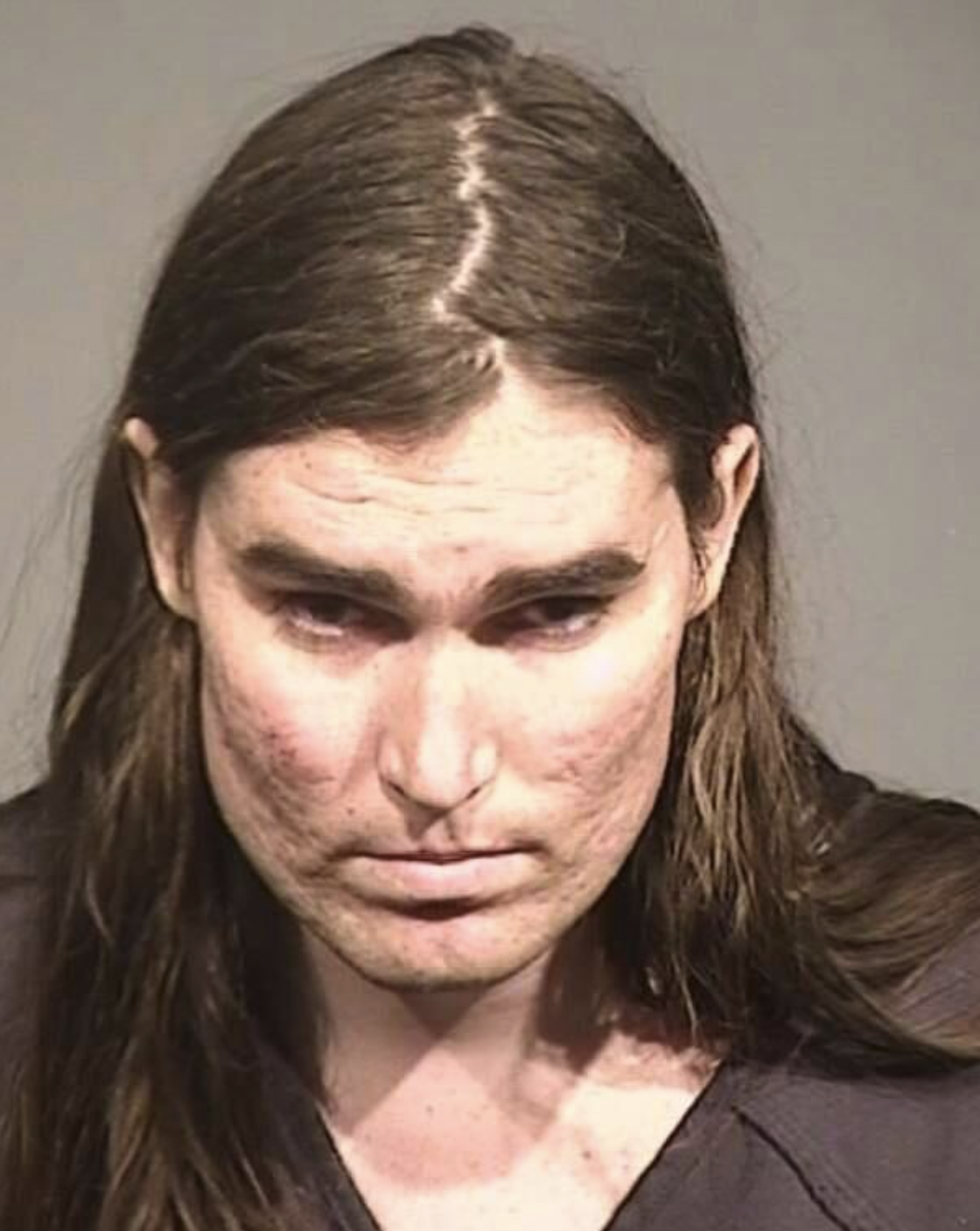 A close‐up booking photograph of an individual with long, straight brown hair parted at the center. They appear to have some acne scarring and are looking downward. The background is neutral gray, and the person is wearing what appears to be a dark shirt or jumpsuit.