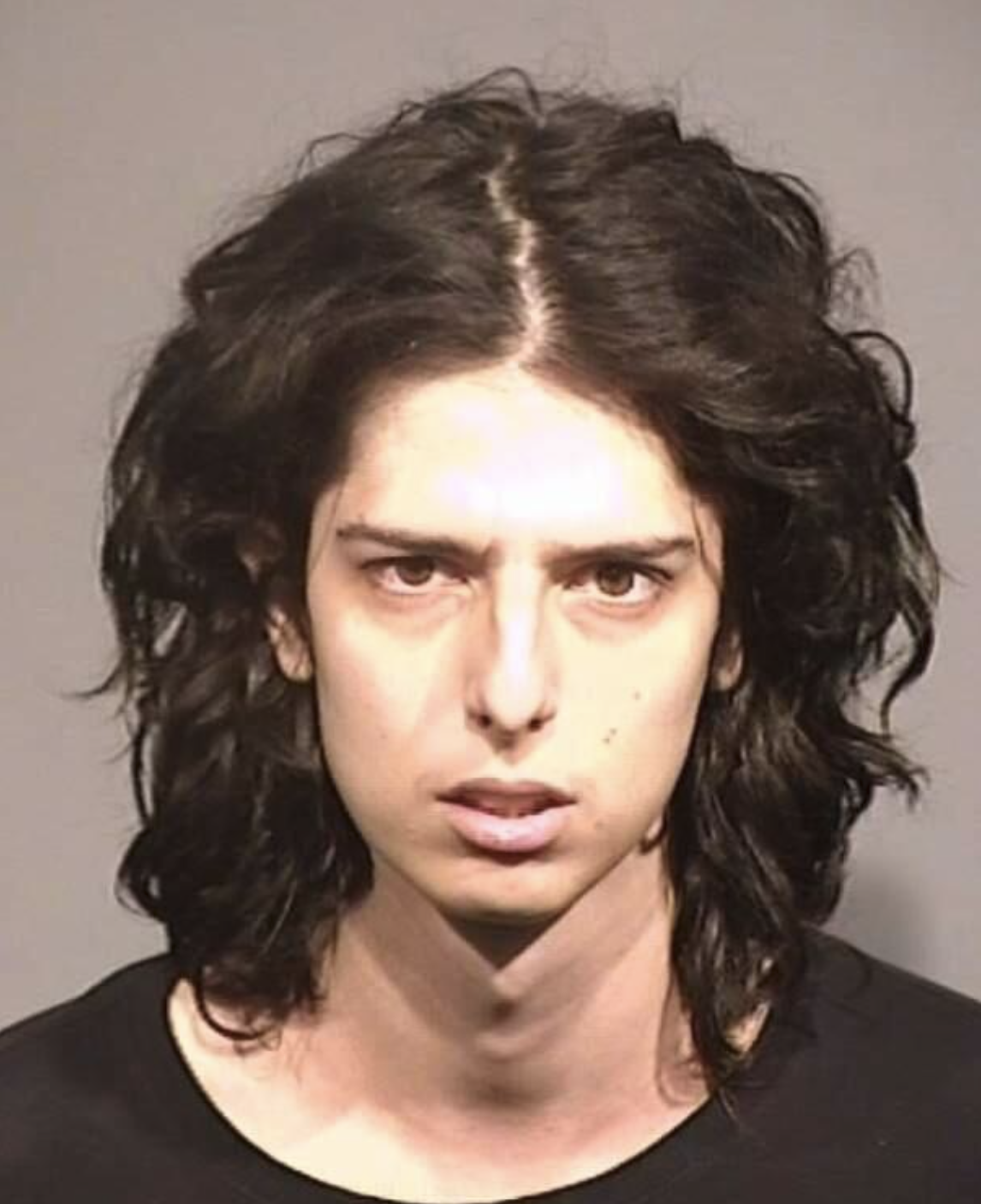 A close‐up booking photograph of an individual with mid‐length, wavy black hair, parted at the center. They are looking directly at the camera with a neutral to serious expression. The background is gray, and the person is wearing a black shirt.