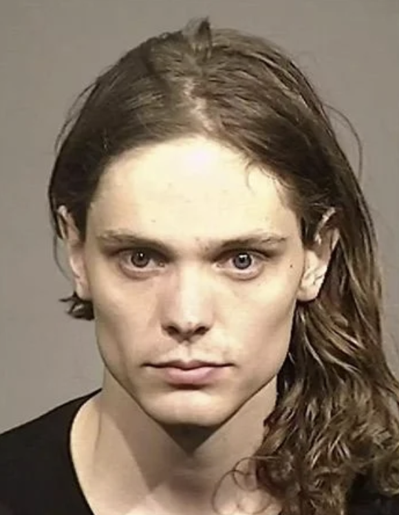 A head-and-shoulders portrait of an adult with long, light-brown hair swept to one side. The person has a neutral expression and looks directly at the camera against a plain, light-gray background. They are wearing a dark, round-neck shirt.