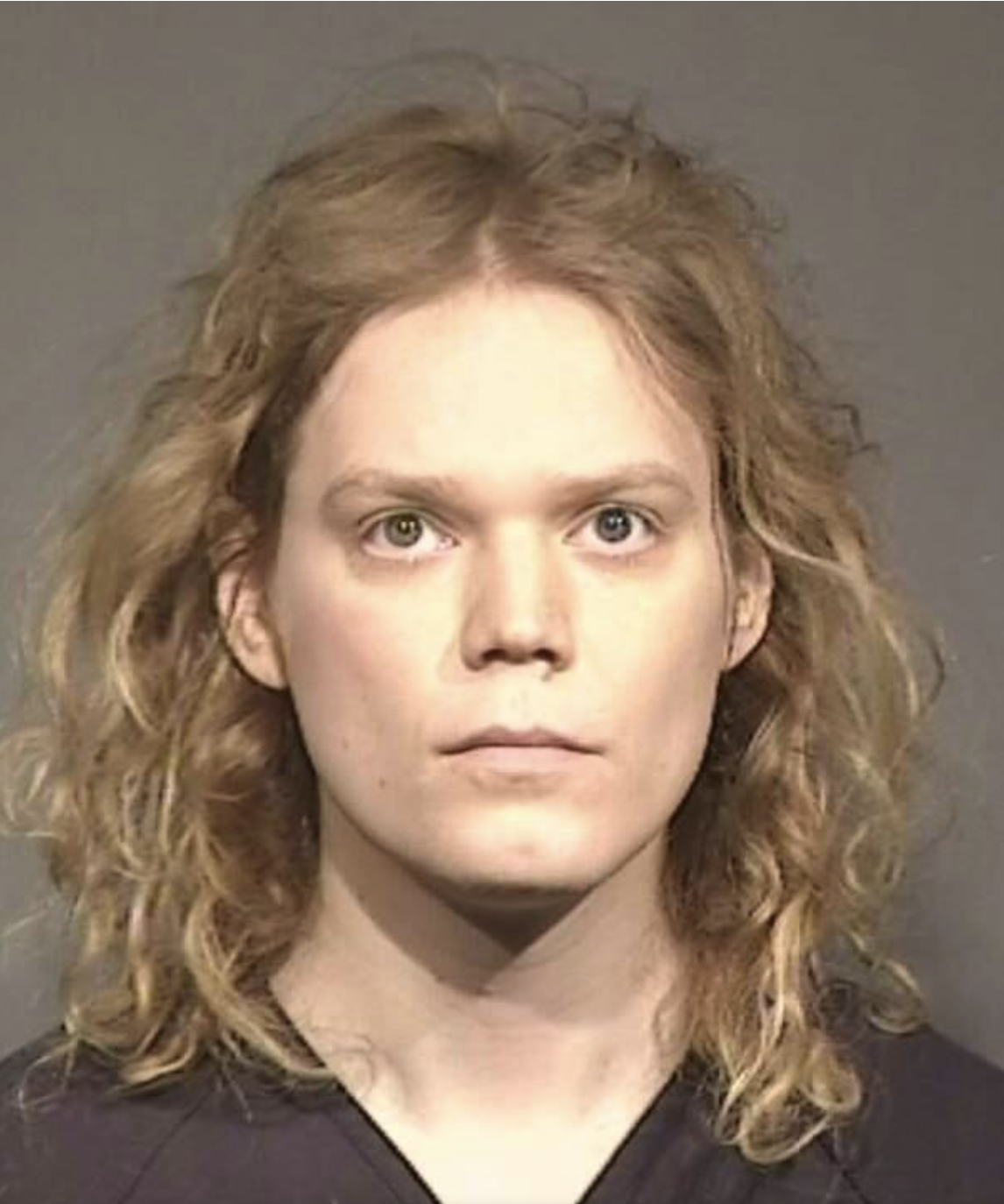 A close‐up booking photograph of an individual with long, wavy blonde hair, parted at the center. The background is neutral gray, and the person is wearing what appears to be a dark shirt or jumpsuit.