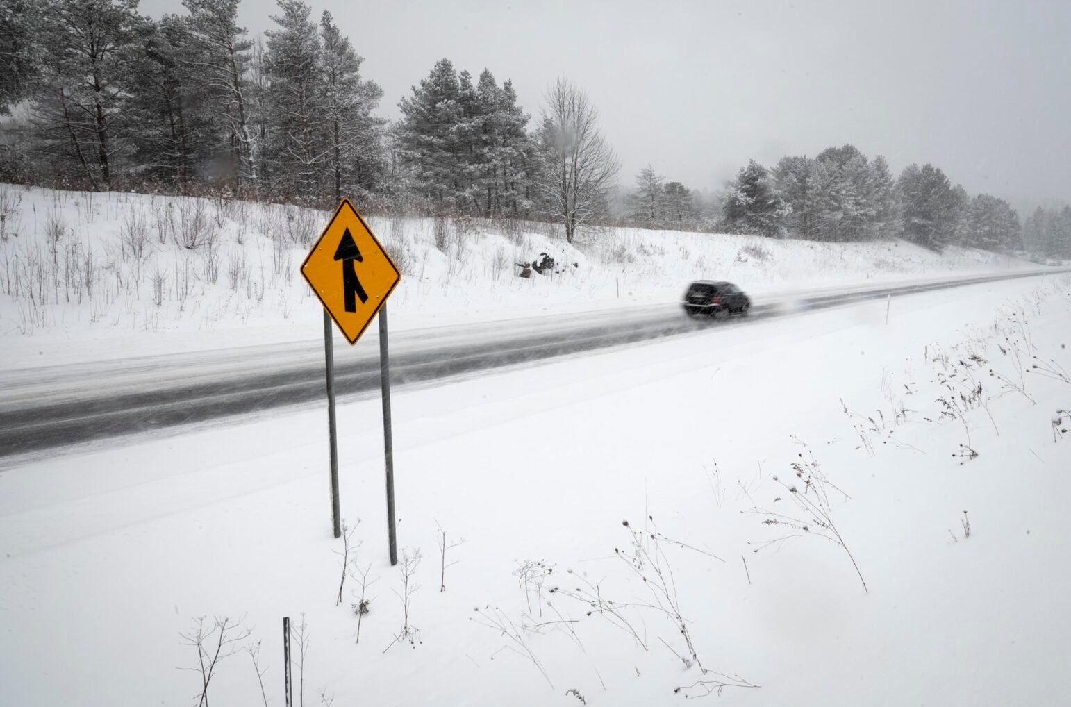 A snow-covered roadway extends into the distance as a black SUV drives past in blurred motion. In the foreground, a bright yellow merge sign stands against a backdrop of white, with leafless shrubs and a tree-lined embankment visible in the background. The scene is enveloped in a wintry haze, with steady snowfall and windblown drifts.