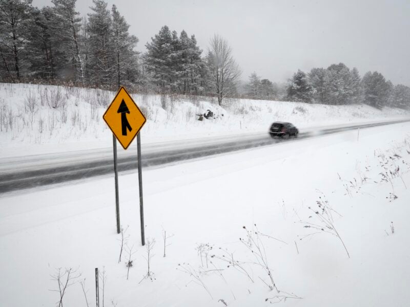 A snow-covered roadway extends into the distance as a black SUV drives past in blurred motion. In the foreground, a bright yellow merge sign stands against a backdrop of white, with leafless shrubs and a tree-lined embankment visible in the background. The scene is enveloped in a wintry haze, with steady snowfall and windblown drifts.