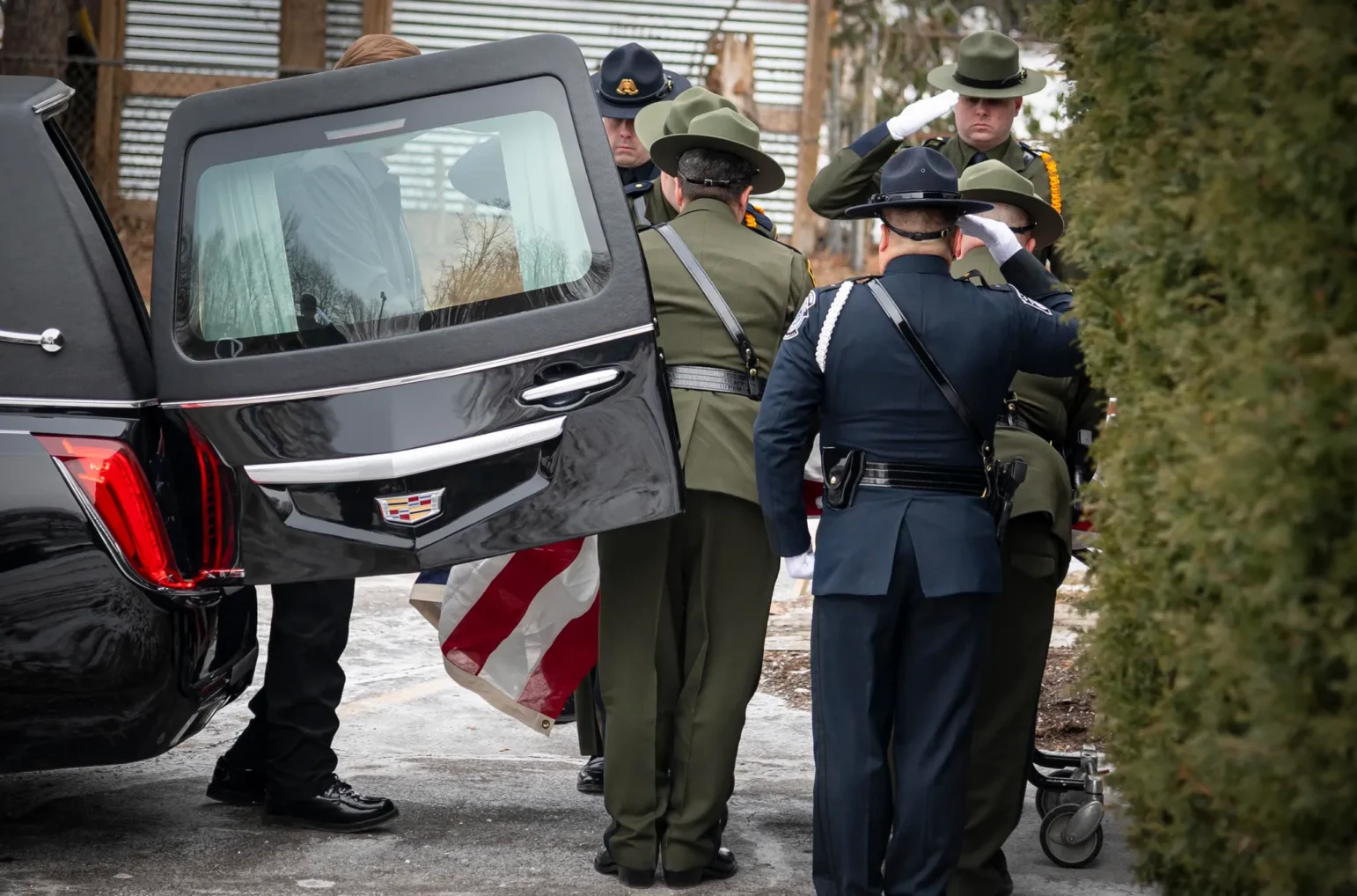 Several uniformed officers stand beside the open rear door of a black Cadillac hearse. They are in formal attire — some wearing campaign hats in green or dark blue — and perform a salute. An American flag-draped casket is partially visible at their feet. The ground around them appears icy, and tall shrubs frame the right edge of the scene.