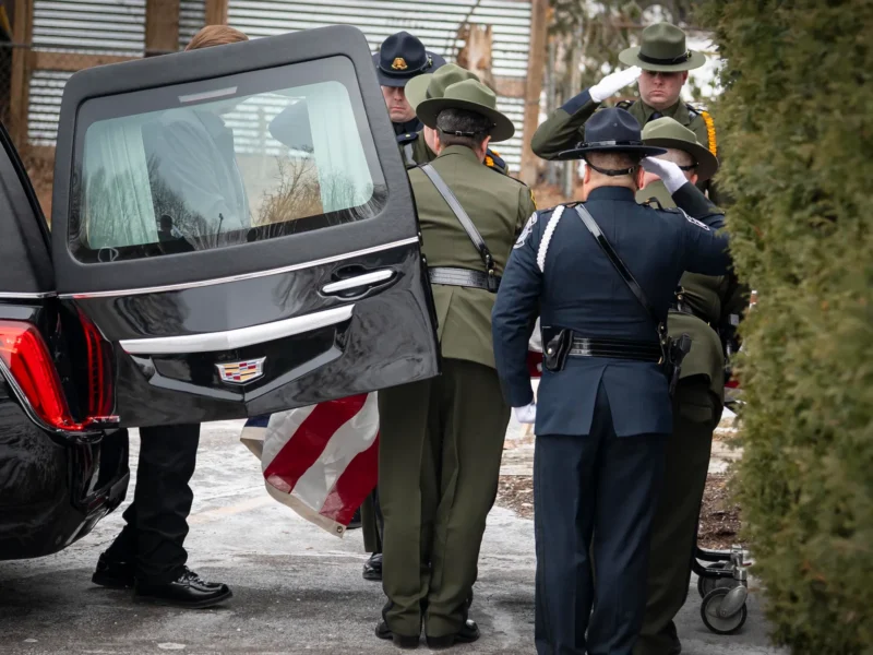 Several uniformed officers stand beside the open rear door of a black Cadillac hearse. They are in formal attire — some wearing campaign hats in green or dark blue — and perform a salute. An American flag-draped casket is partially visible at their feet. The ground around them appears icy, and tall shrubs frame the right edge of the scene.