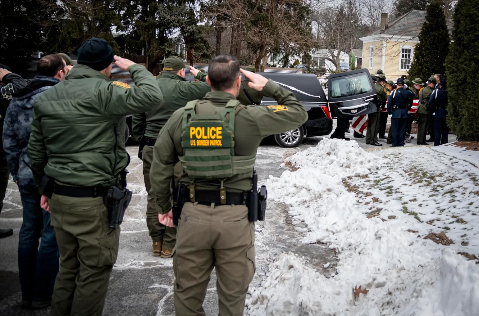 Several uniformed U.S. Border Patrol officers, dressed in green jackets and vests bearing “POLICE U.S. Border Patrol,” stand outdoors in a snowy area. They are arranged in a line with their right hands raised in salute as a black hearse, carrying a flag‐draped casket, passes by. A yellow house and trees are visible in the background.