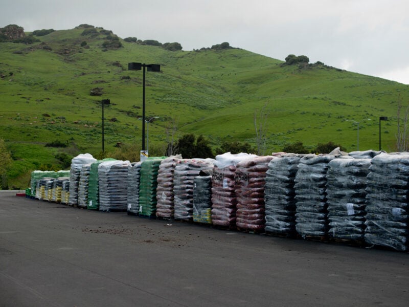 Stacks of bagged landscaping materials, wrapped in plastic, are neatly lined up in an outdoor parking lot. The colorful bags contrast with the lush green rolling hills and cloudy sky in the background, creating a blend of industrial and natural landscapes.