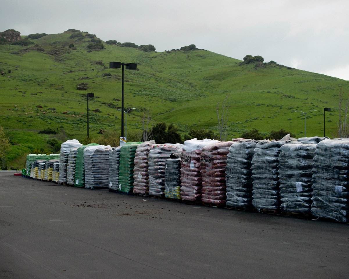 Stacks of bagged landscaping materials, wrapped in plastic, are neatly lined up in an outdoor parking lot. The colorful bags contrast with the lush green rolling hills and cloudy sky in the background, creating a blend of industrial and natural landscapes.