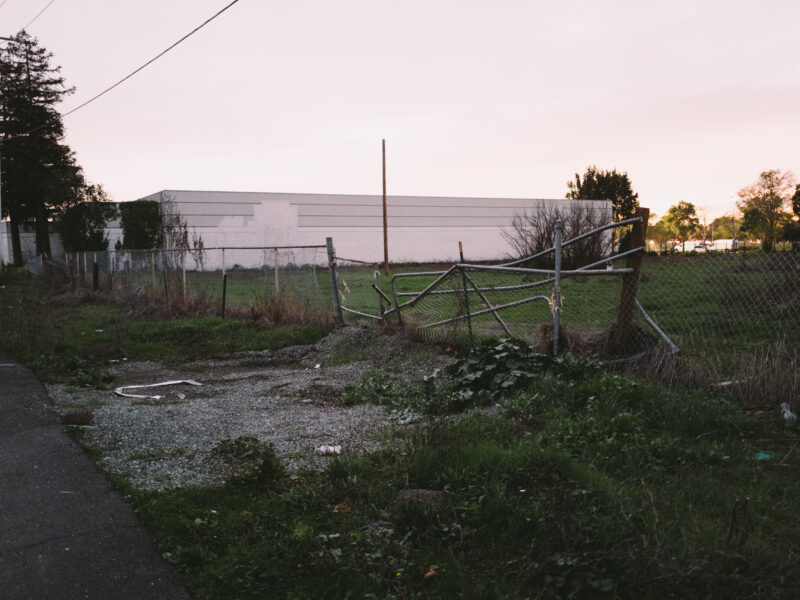 A chain-link fence along a narrow grassy lot is partially collapsed, its metal gate bent near a gravel patch. A tall, white rectangular building stands behind the fenced area under a pale sky at dusk. Power lines and a few tall coniferous trees line the left edge of the scene, while scattered debris and overgrown vegetation cover the ground in the foreground.