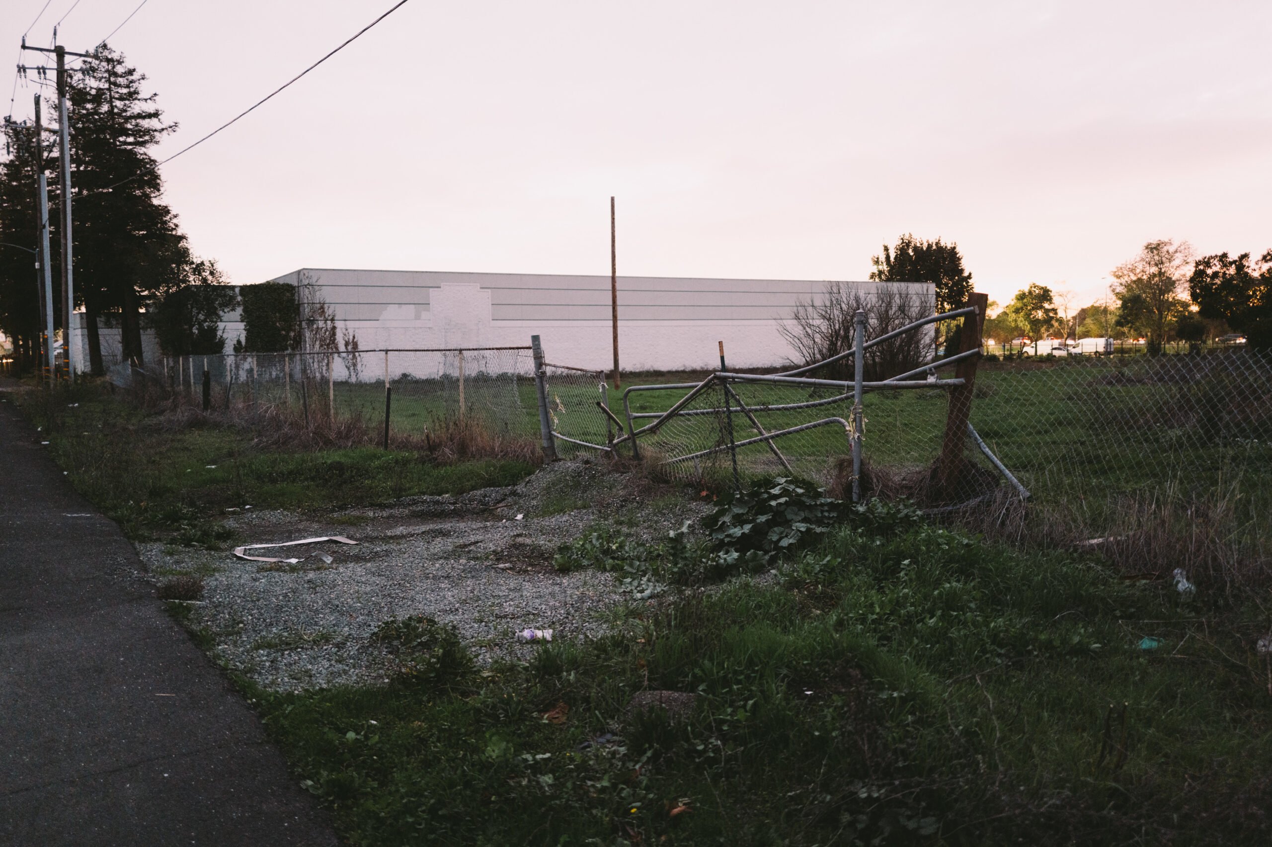 A chain-link fence along a narrow grassy lot is partially collapsed, its metal gate bent near a gravel patch. A tall, white rectangular building stands behind the fenced area under a pale sky at dusk. Power lines and a few tall coniferous trees line the left edge of the scene, while scattered debris and overgrown vegetation cover the ground in the foreground.