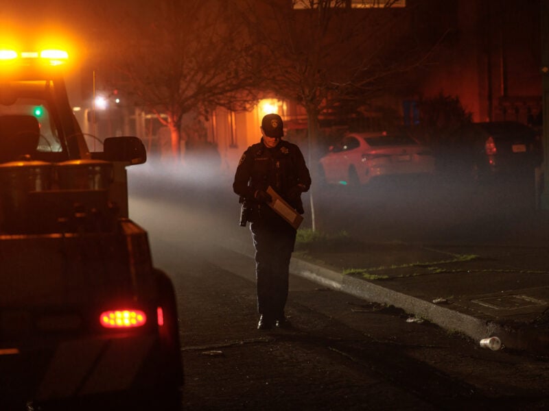 A police officer in dark uniform walks along a dimly lit residential street carrying a clipboard or folder. The area is shrouded in haze or smoke, illuminated by the orange emergency lights of a tow truck parked nearby. Several cars and a bare tree line the background, with warm lights from houses visible behind a soft fog.