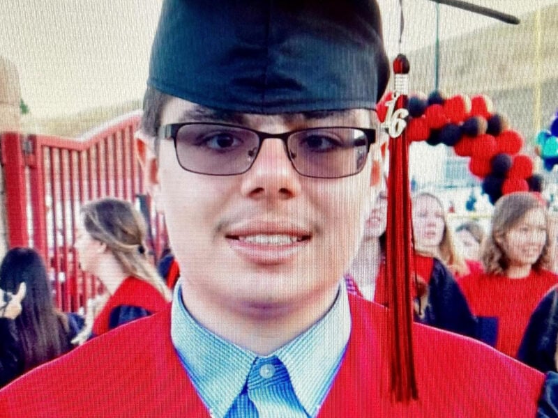 A young adult wearing glasses and a black graduation cap with a tassel reading “16” stands in front of a crowd at a commencement ceremony. He’s dressed in a robe with a red stole, over a collared button-down shirt, and holds a black diploma cover bearing the seal of the San Ramon Valley Unified School District. A red and black balloon arch and other graduates are visible in the background.