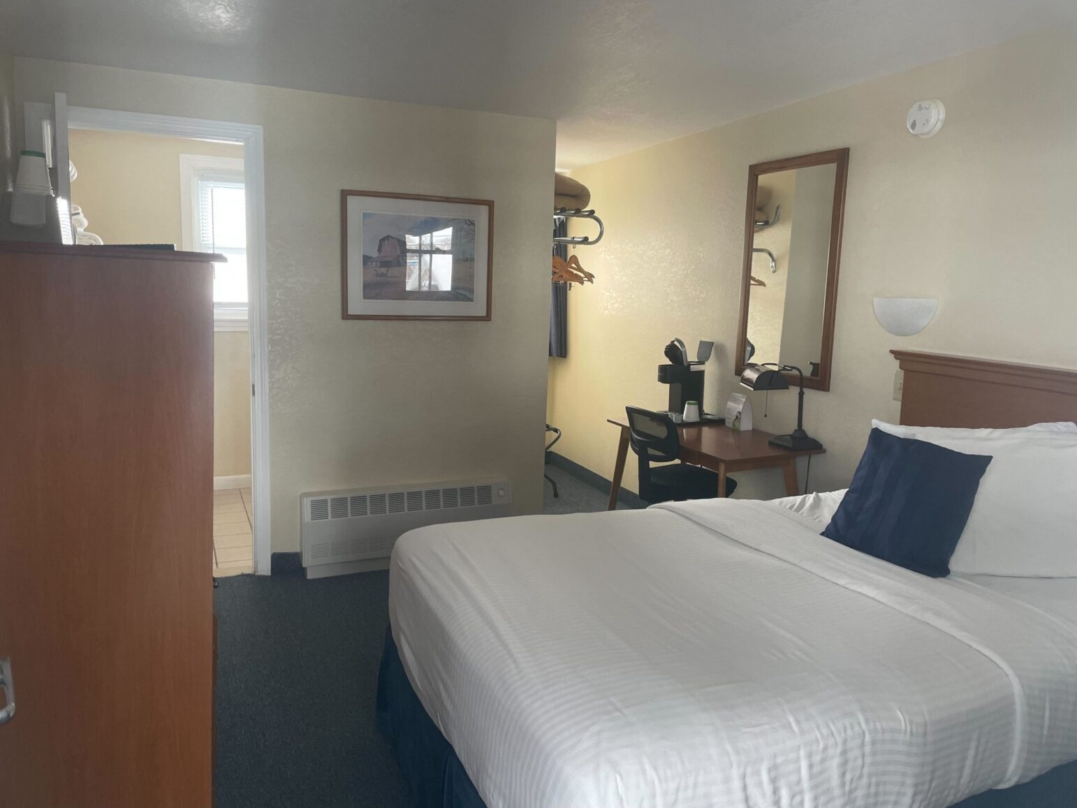 A small hotel room with cream-colored walls and a neatly made bed featuring white linens and a dark blue accent pillow. A wooden wardrobe and a small desk with a coffee machine are positioned along one wall. The open door to a tiled bathroom is visible in the left background, allowing daylight to enter.