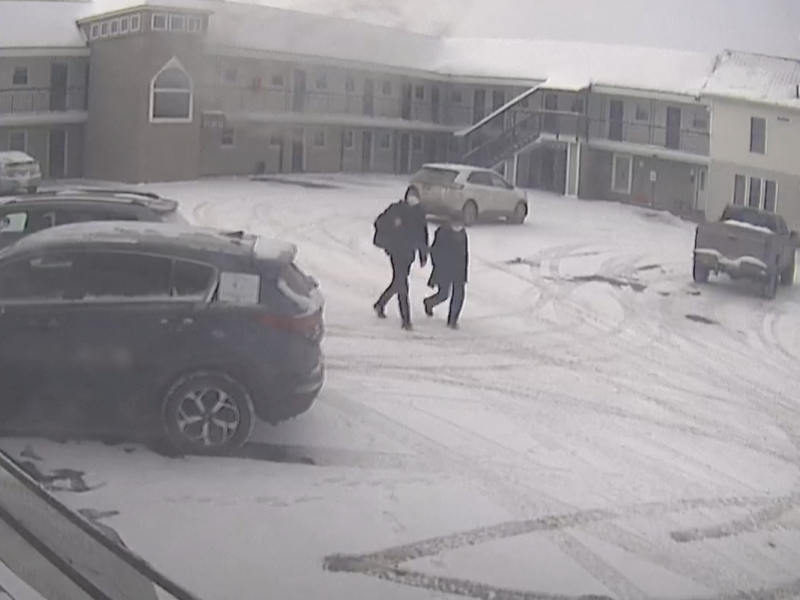 A grainy security camera view of a two-story motel courtyard in winter. Several cars, lightly dusted with snow, are parked on a snowy lot. In the center of the frame, two people in dark jackets walk side by side across the parking area. A staircase and balconies are visible on the motel’s upper level.