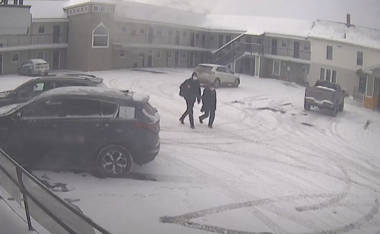 A grainy security camera view of a two-story motel courtyard in winter. Several cars, lightly dusted with snow, are parked on a snowy lot. In the center of the frame, two people in dark jackets walk side by side across the parking area. A staircase and balconies are visible on the motel’s upper level.