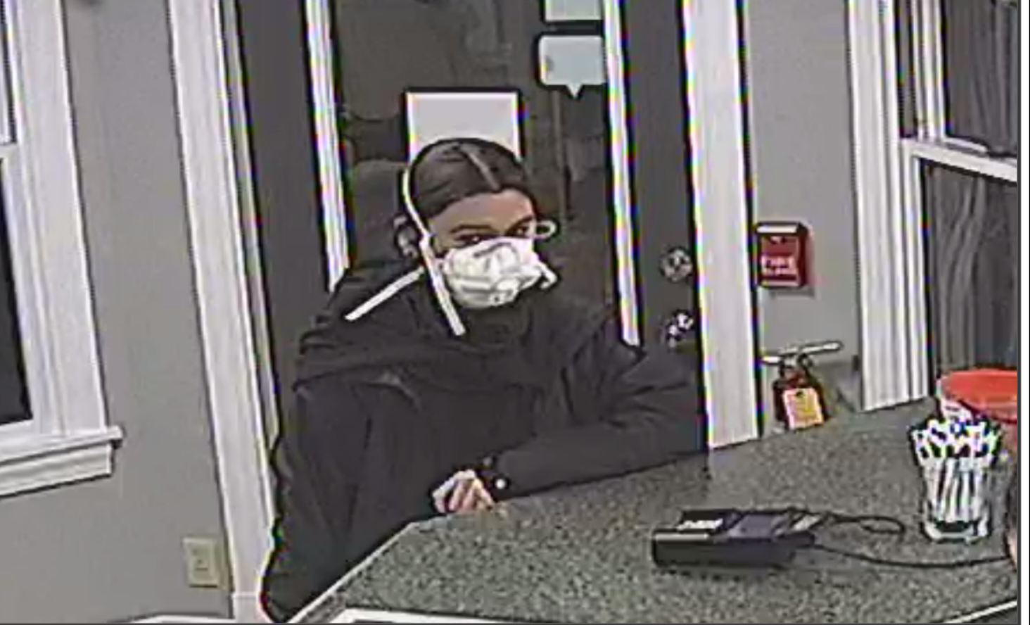 A low-resolution security camera image shows a person with dark, center-parted hair wearing a white, cup-style respirator mask and a dark jacket or hoodie. They lean on a countertop inside what appears to be a reception area, with a door and windows behind them.