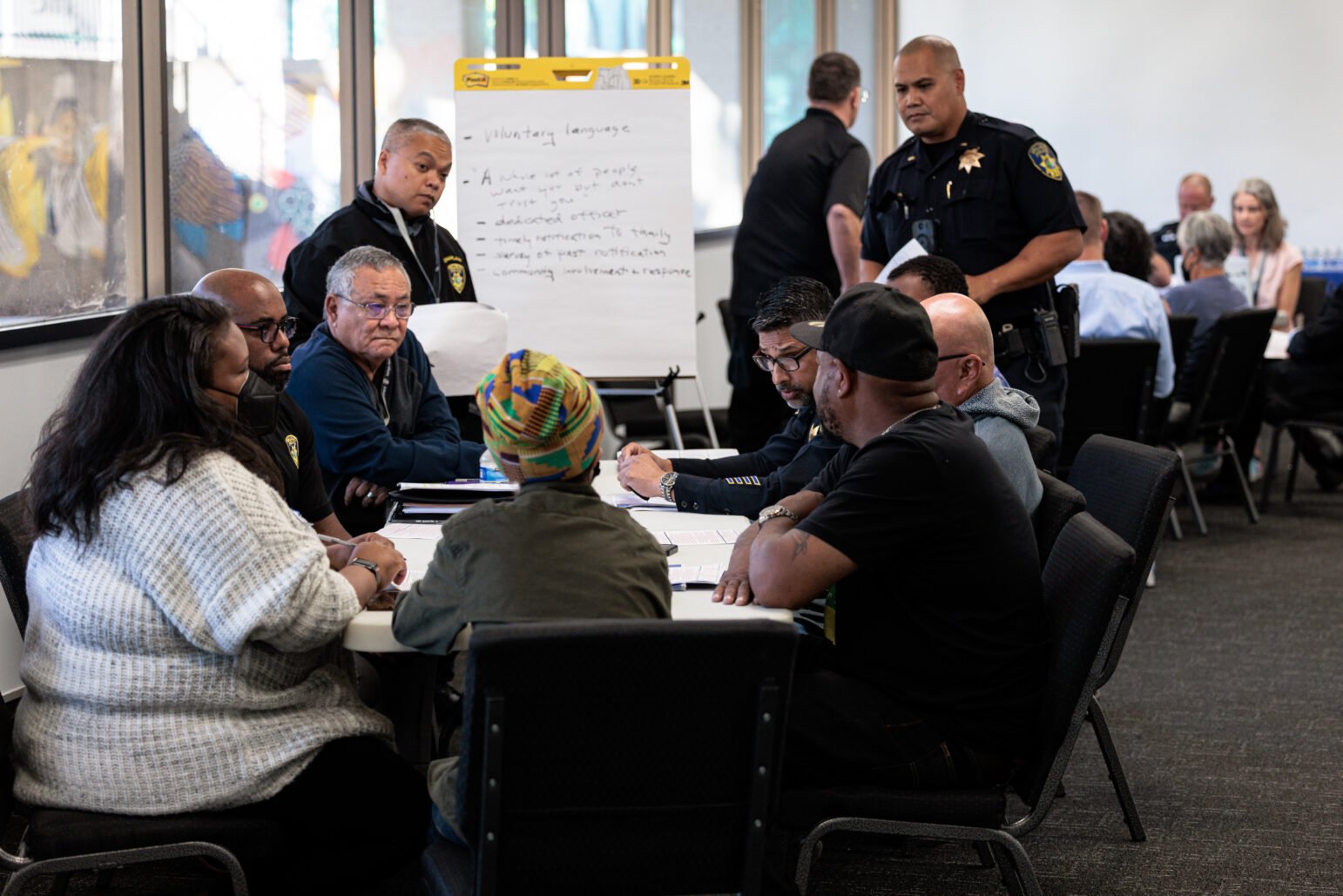 A group of people, including several uniformed police officers, sit around a table in a large, well-lit meeting room. A flipchart with handwritten notes stands behind them, and other participants are gathered at tables in the background. One individual wears a colorful patterned headwrap, while another person in a knit sweater faces the group. A few officers stand nearby, observing or listening to the discussion.