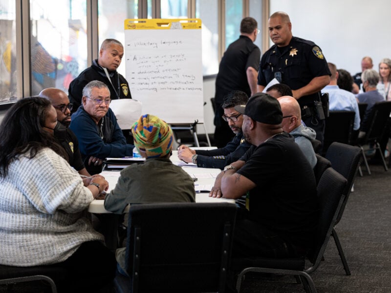 A group of people, including several uniformed police officers, sit around a table in a large, well-lit meeting room. A flipchart with handwritten notes stands behind them, and other participants are gathered at tables in the background. One individual wears a colorful patterned headwrap, while another person in a knit sweater faces the group. A few officers stand nearby, observing or listening to the discussion.