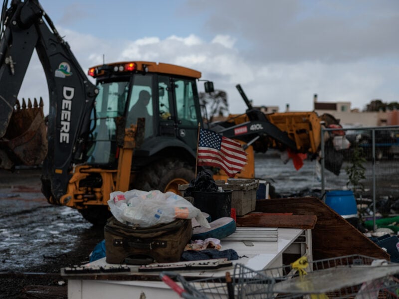 A muddy lot with a large yellow City of Vallejo backhoe parked in the center. In the foreground, a small, weathered American flag stands among scattered belongings including plastic bags, a suitcase, and wooden debris.