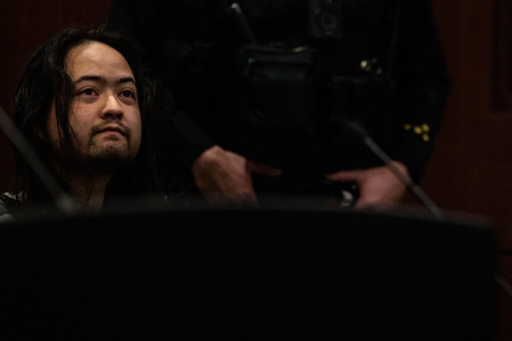 A man sits quietly in a courtroom, appearing reflective and looking upward, with a partially visible uniformed officer standing behind him. The setting is somber with dim lighting, suggesting a judicial proceeding.