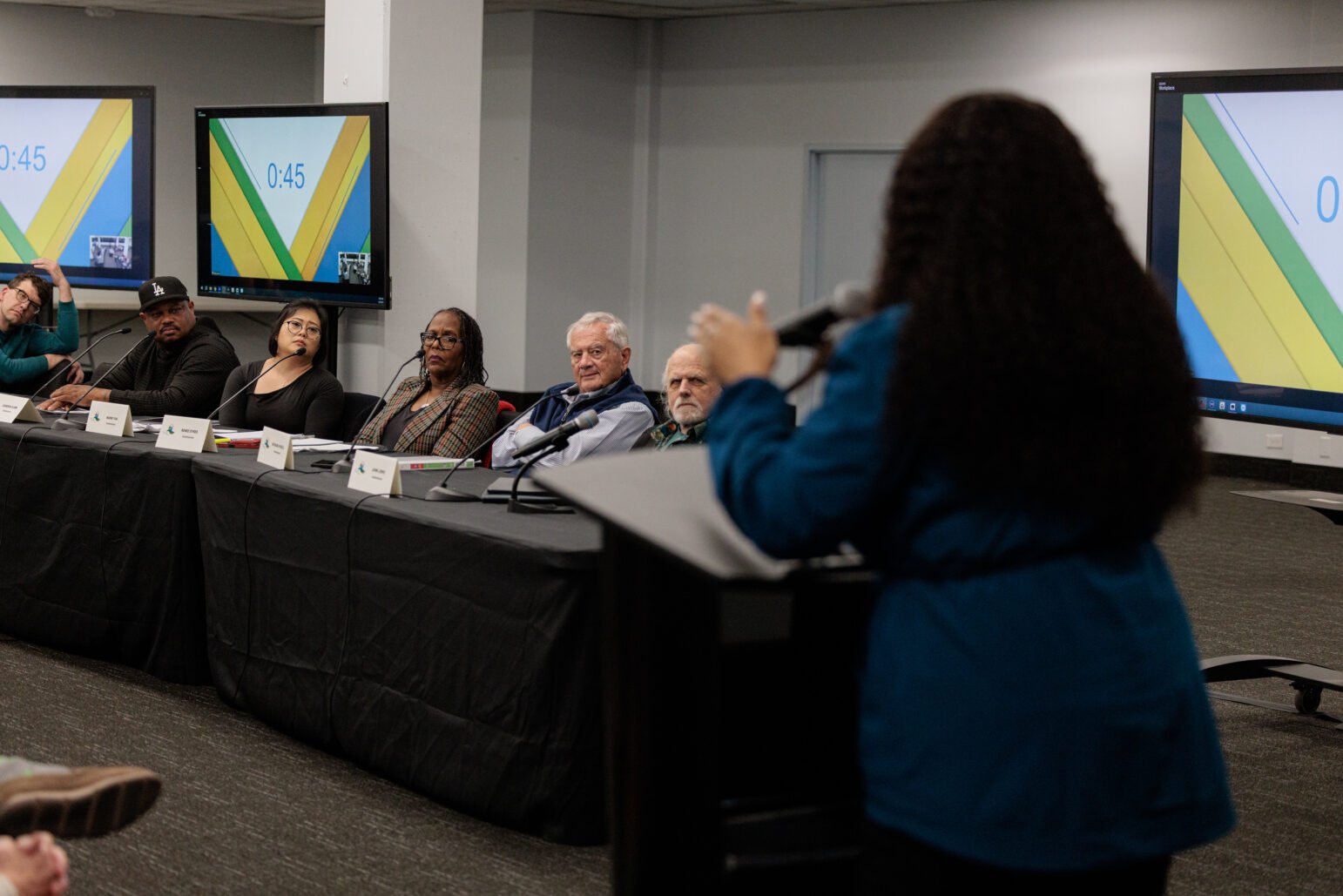 A woman with long, curly hair stands at a podium, speaking to a panel of six individuals seated behind a black-clothed table. Large monitors behind them display a digital timer (“0:45”) against green and yellow background graphics. The panelists, each with a name card in front of them, listen attentively.