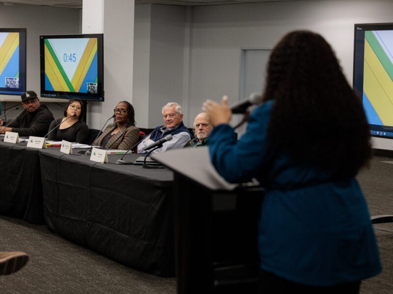 A woman with long, curly hair stands at a podium, speaking to a panel of six individuals seated behind a black-clothed table. Large monitors behind them display a digital timer (“0:45”) against green and yellow background graphics. The panelists, each with a name card in front of them, listen attentively.