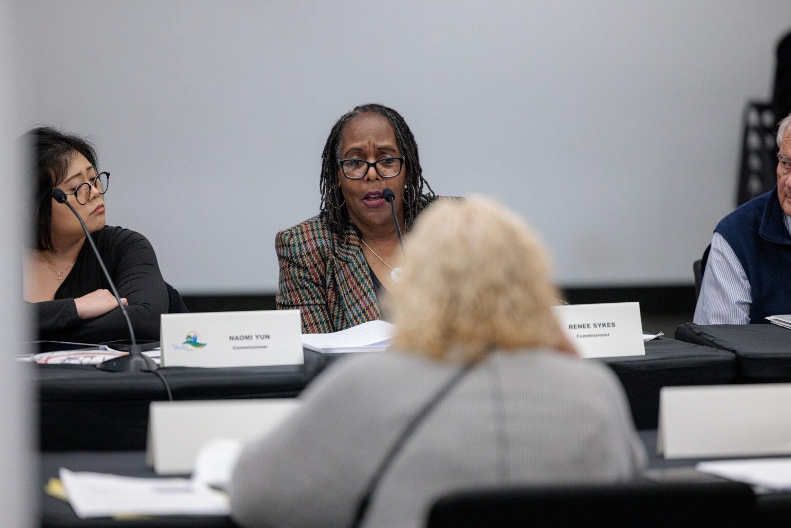 A closer view of the same panel. The commissioner in the center, wearing a plaid blazer and glasses, speaks into a microphone. Her nameplate reads “Renee Sykes, Commissioner.” To her left, another commissioner with short hair and glasses looks on (labelled “Naomi Yun, Commissioner”). An older male panelist is partially visible on the right. A figure in the foreground faces the panel, slightly out of focus.