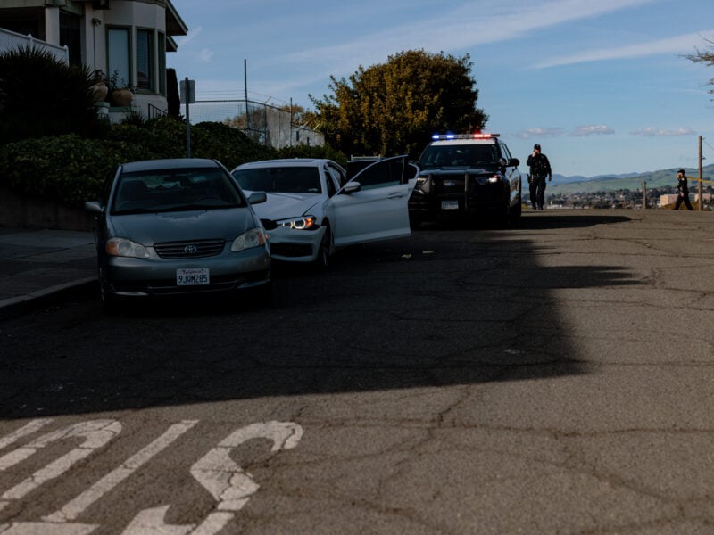 A police officer walks toward a parked patrol vehicle with emergency lights activated on a quiet residential street, while another officer in the background unfurls crime scene tape. Nearby, a white sedan sits with its driver-side door open, stopped behind a gray Toyota sedan. The street is calm under clear skies.