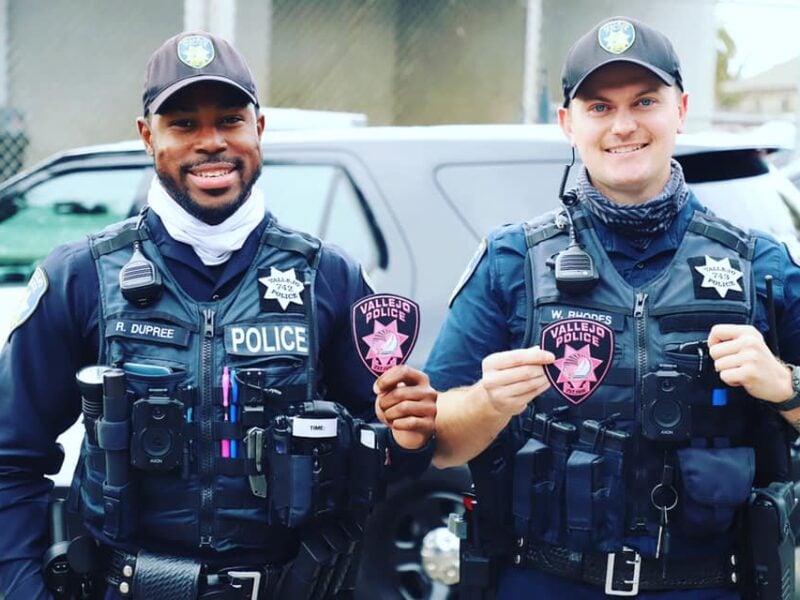 Two uniformed police officers stand side by side, both wearing dark ballistic vests with “POLICE” markings and matching black baseball caps bearing a department badge. Each officer holds a pink patch with “Vallejo Police” and a stylized star, presumably to support breast cancer awareness. A black police SUV is parked behind them. Both officers are smiling as they pose for the photo.