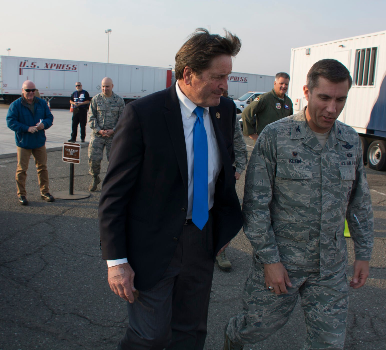 A man in a dark suit and bright blue tie walks beside a U.S. Air Force officer in camouflage on an asphalt surface, with other personnel and trailers in the background.