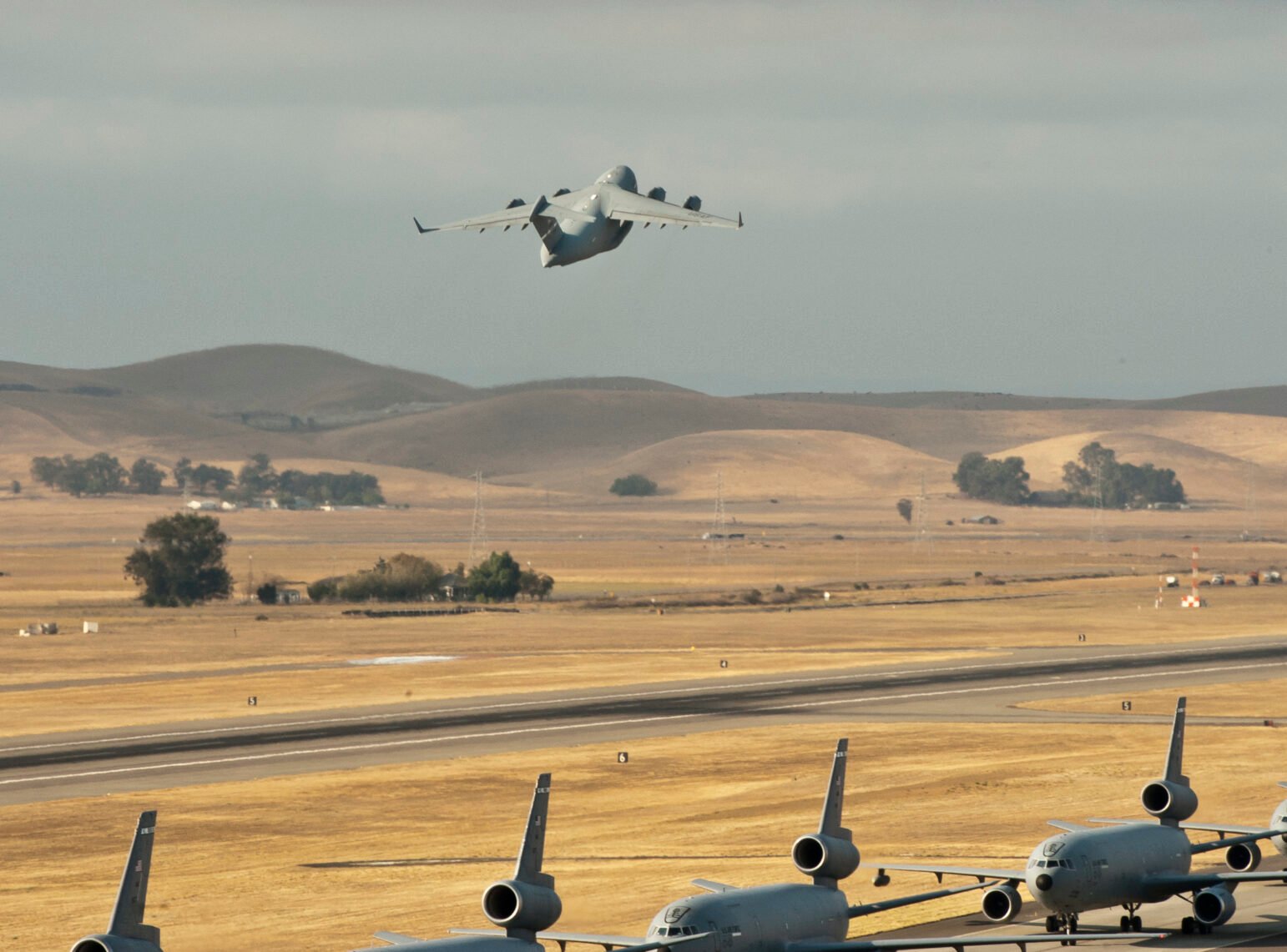 A large military transport jet climbs over dry, rolling hills as a row of gray Air Force aircraft waits on the runway below.