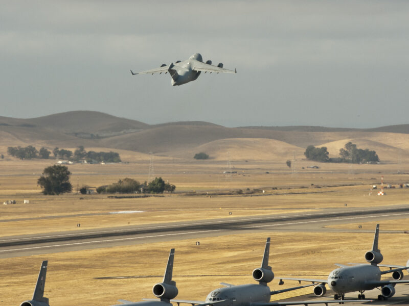 A large military transport jet climbs over dry, rolling hills as a row of gray Air Force aircraft waits on the runway below.