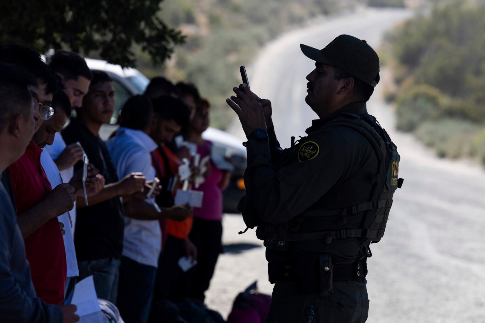 A U.S. Border Patrol agent in dark uniform photographs a line of detained migrants holding documents along a remote desert roadside.