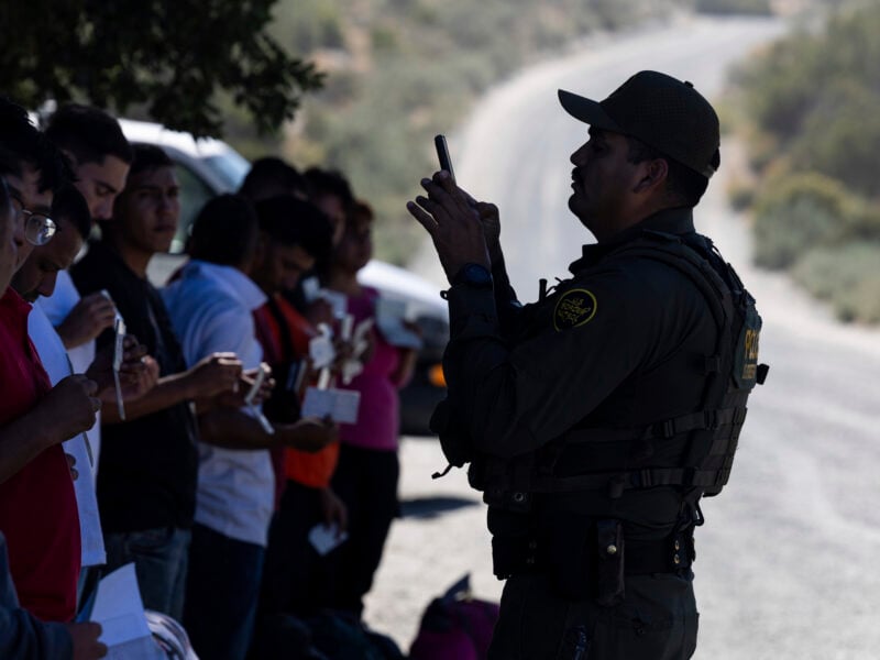 A U.S. Border Patrol agent in dark uniform photographs a line of detained migrants holding documents along a remote desert roadside.