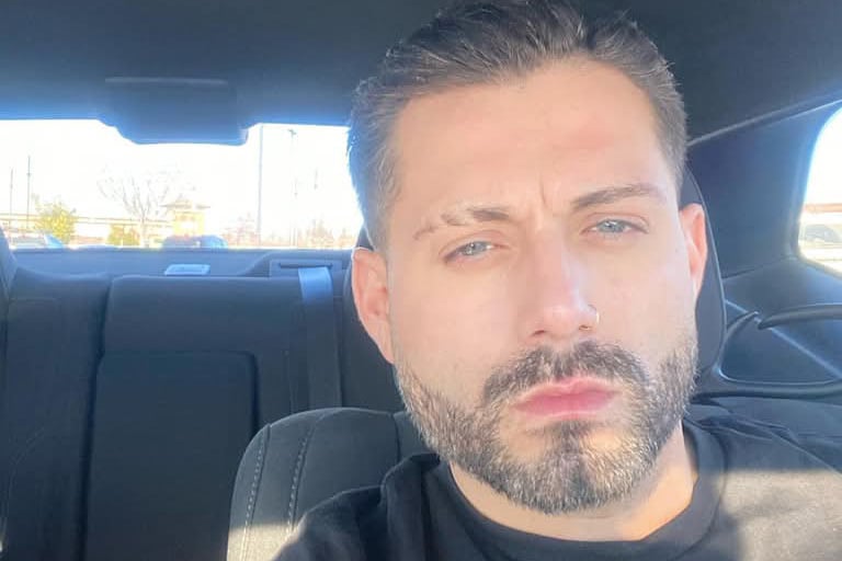 Man with close-cropped dark hair and trimmed beard sits in a car, facing the camera in bright daylight.