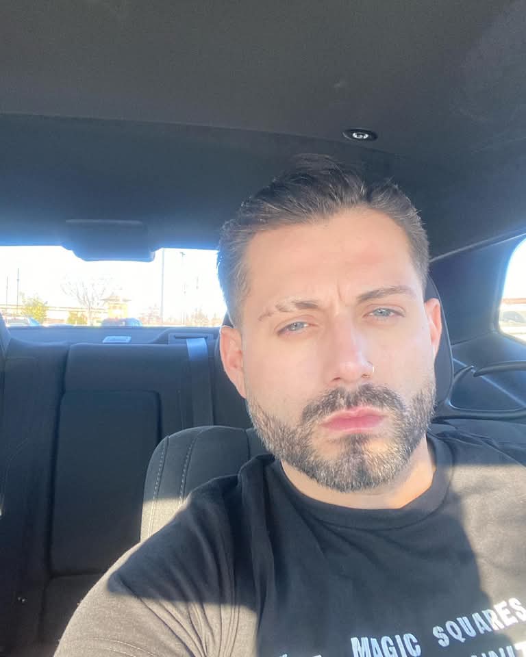 Man with close-cropped dark hair and trimmed beard sits in a car, facing the camera in bright daylight.