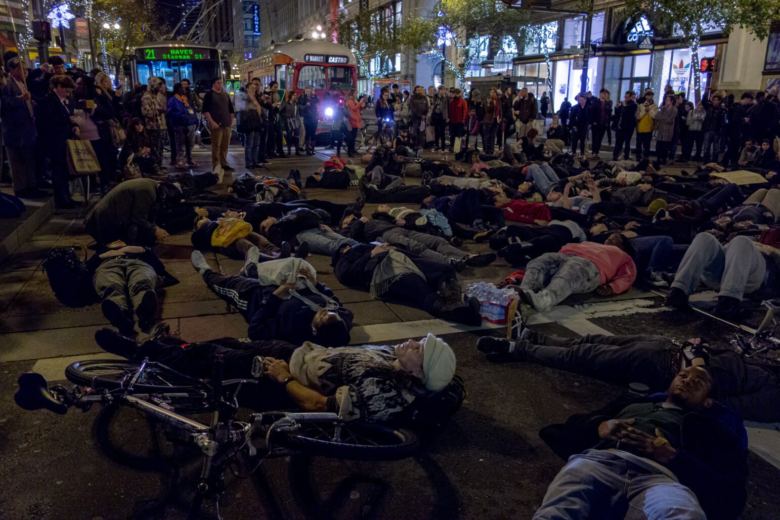 A group of demonstrators lie down on a city street at night during a "die-in" protest against police violence. Spectators watch from the sidewalk, and city public transit is halted by the protest.