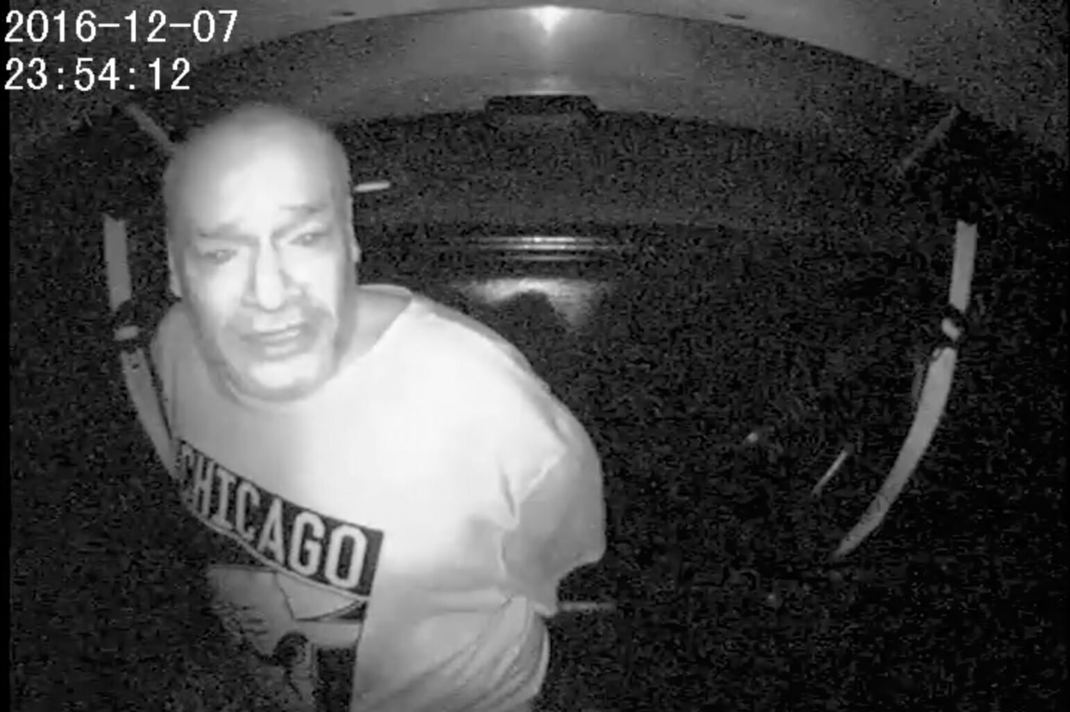 Grainy black-and-white surveillance image shows Darryl Mefferd, handcuffed and wearing a Chicago Bulls shirt, leaning forward in the backseat of a police vehicle. He appears distressed. A timestamp in the upper-left corner reads “2016-12-07 23:54:12.”