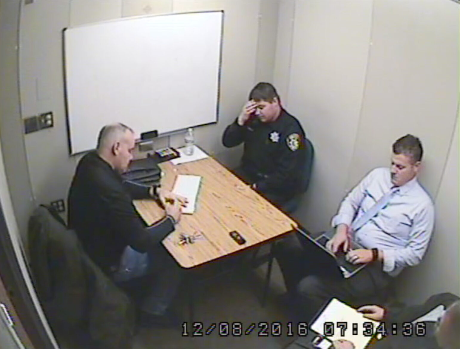 Surveillance camera image showing Vallejo Police Ofc. Jeremy Callinan sitting at a table in a small, brightly-lit interrogation room. He appears stressed, with his hand covering part of his face. Two men sit near him, one taking handwritten notes, and another using a laptop.