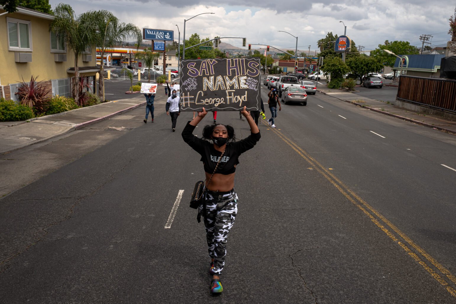 A demonstrator marches down a wide, mostly empty street holding a sign overhead that reads "Say His Name #GeorgeFloyd." Other protesters follow behind in the distance.