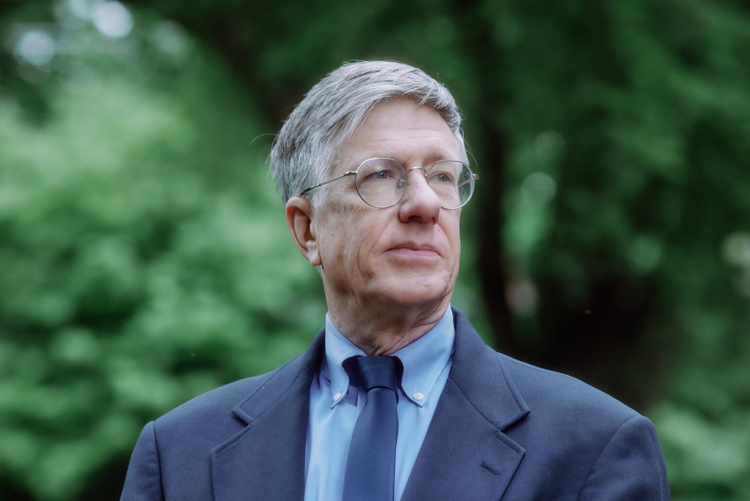 Portrait of Dr. Victor Weedn standing outdoors, wearing glasses and a formal dark suit. He has a neutral expression and gazes off to the side against a background of lush greenery.